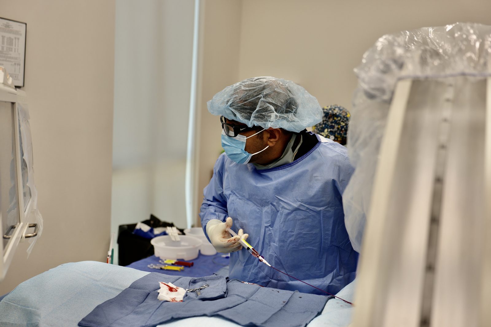 Dr. Samy Al-Bayati, gowned, masked and gloved, holding a catheter and syringe during a hemorrhoid artery embolization in the ARV Centers angiography suite