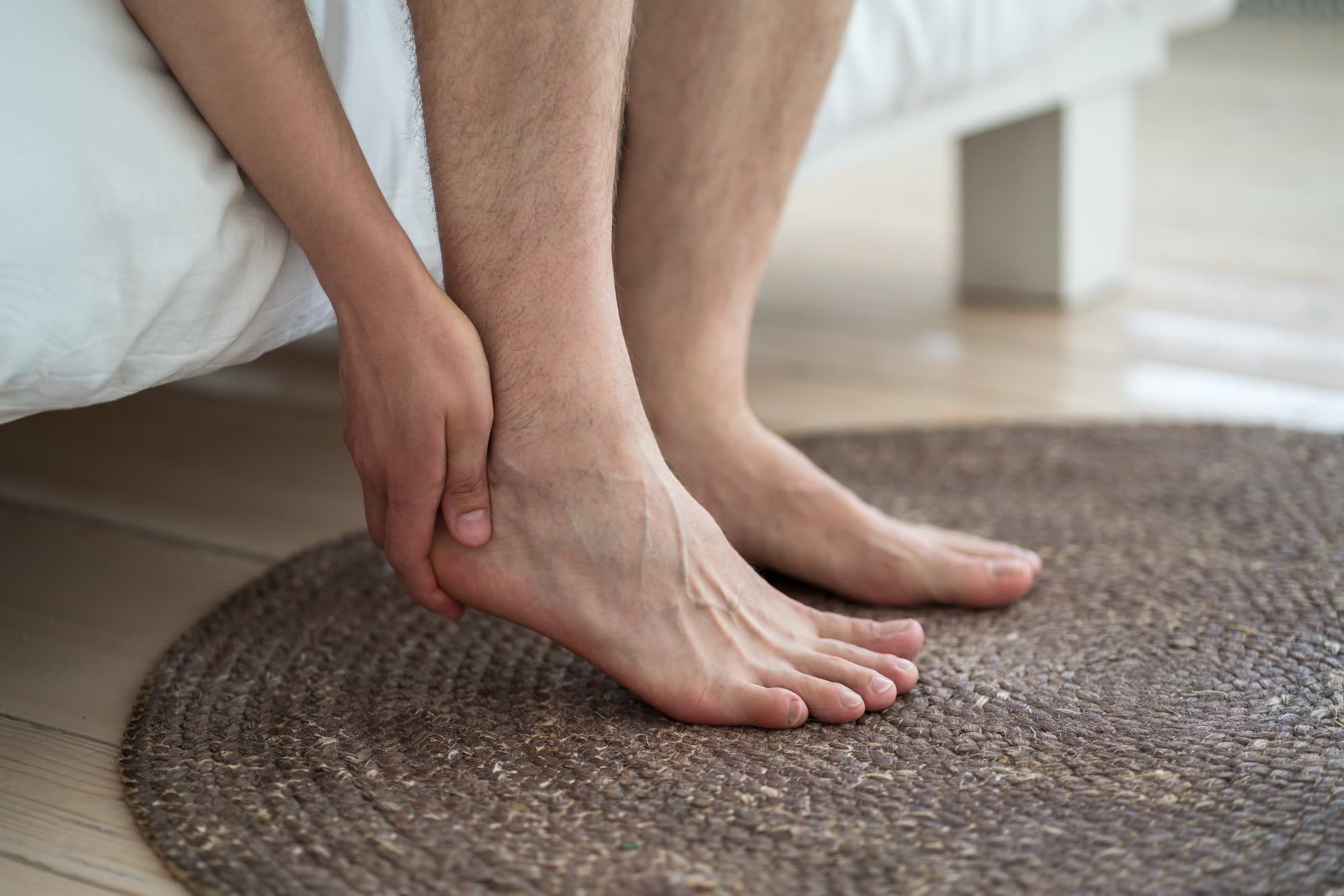 A person sitting on the edge of a bed in the morning, one hand pressed to the heel before standing up