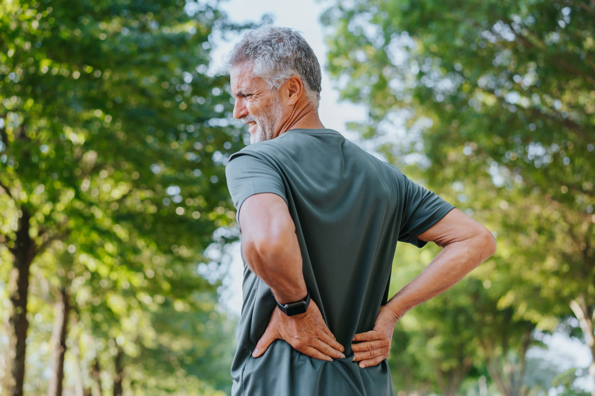 A gray-haired man outdoors presses a hand to one side of his low back, just below the belt line