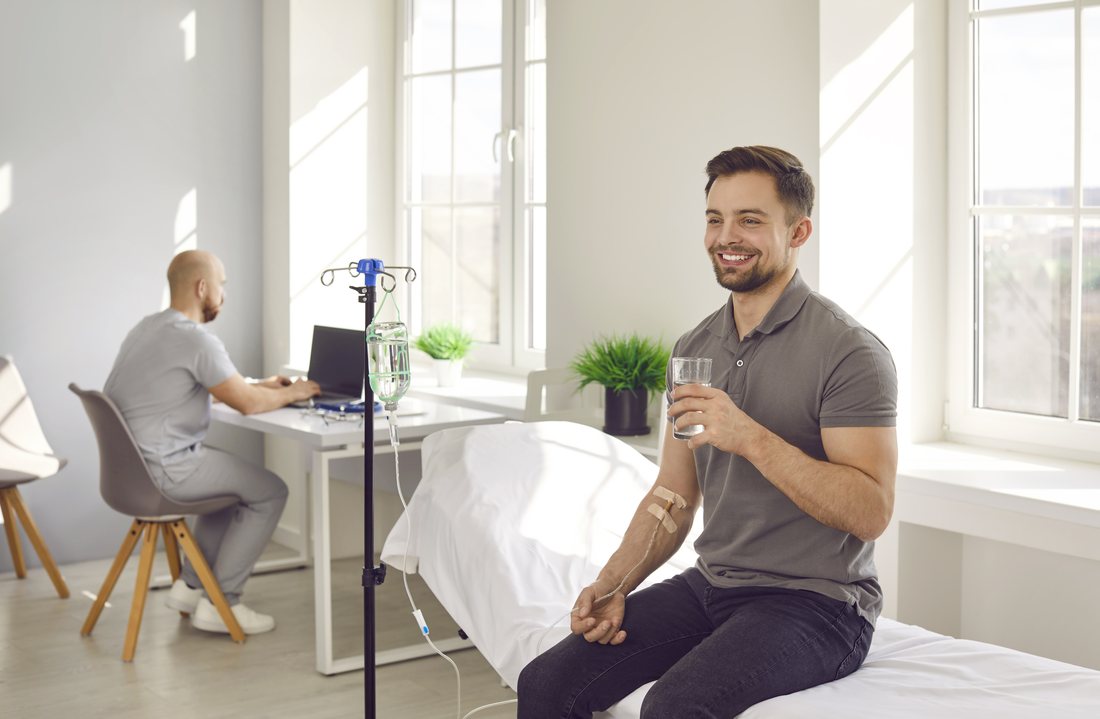 A man sitting up on a clinic recovery bed with an IV line, holding a glass of water and smiling