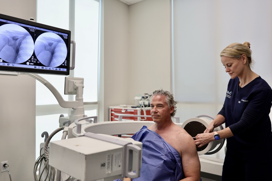 Dr. Lisa Persyn performing a shoulder injection under live fluoroscopic guidance, with the joint visible on the imaging monitor