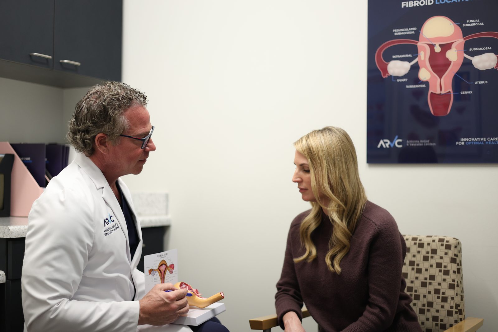 A physician at ARV Centers talking through fibroid treatment options with a uterus model in hand, beneath a poster showing where fibroids grow in and around the uterus